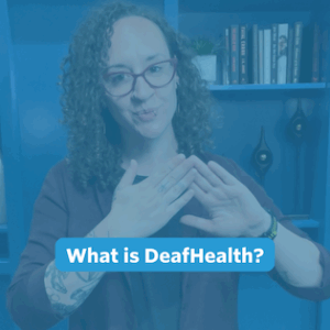 A nonbinary person with long, curly brown hair stands in front of the camera. Behind them is a blue built-in bookshelf wall. They are wearing black pants, a black shirt layered with a maroon button-down shirt, and a pair of gold hexagon earrings.