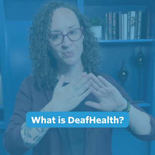 A nonbinary person with long, curly brown hair stands in front of the camera. Behind them is a blue built-in bookshelf wall. They are wearing black pants, a black shirt layered with a maroon button-down shirt, and a pair of gold hexagon earrings.